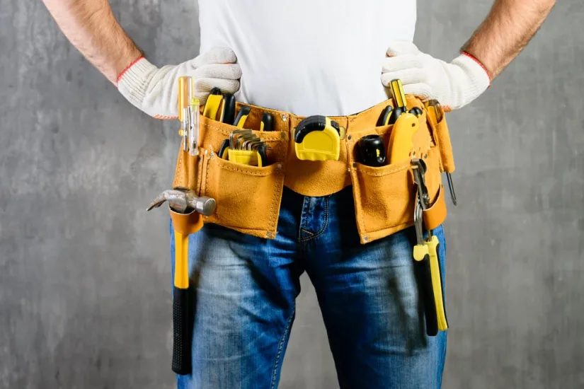 A handyman with hands on waist and tool belt with construction tools against grey background